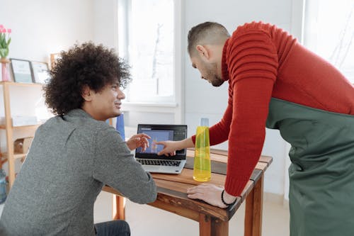 Man in Red Sweater and Green Apron Pointing at the Laptop Man in Gray Shirt Using