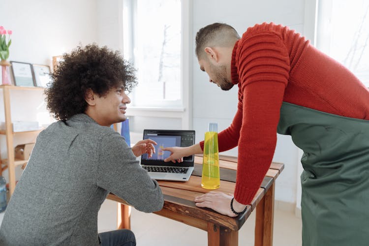 Man In Red Sweater And Green Apron Pointing At The Laptop Man In Gray Shirt Using