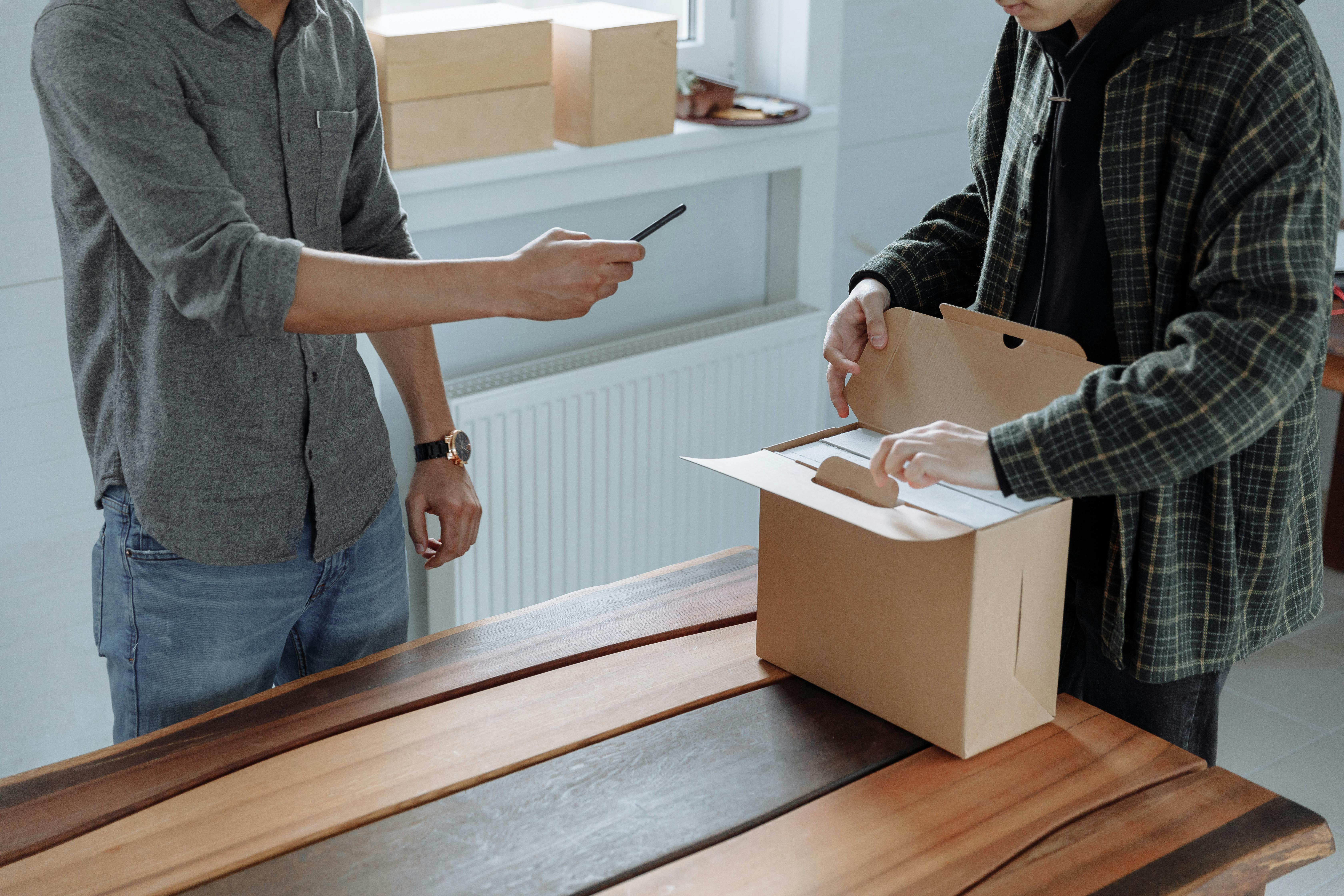 Two adults unpack and photograph a cardboard box using a smartphone indoors.