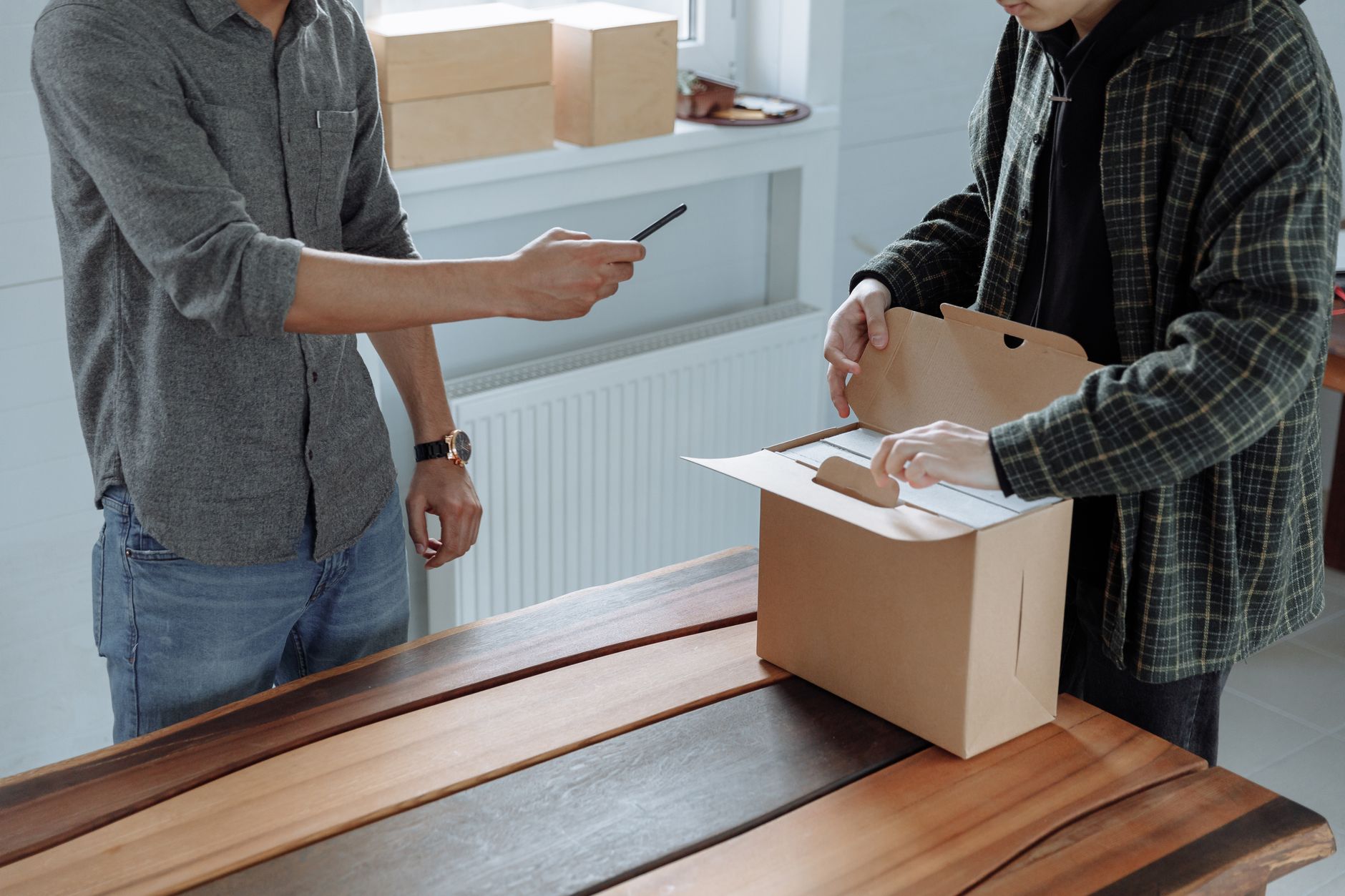 Two adults unpack and photograph a cardboard box using a smartphone indoors.