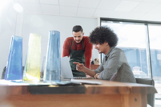 Two colleagues work together on a laptop in a bright office space, surrounded by colorful glass vases.