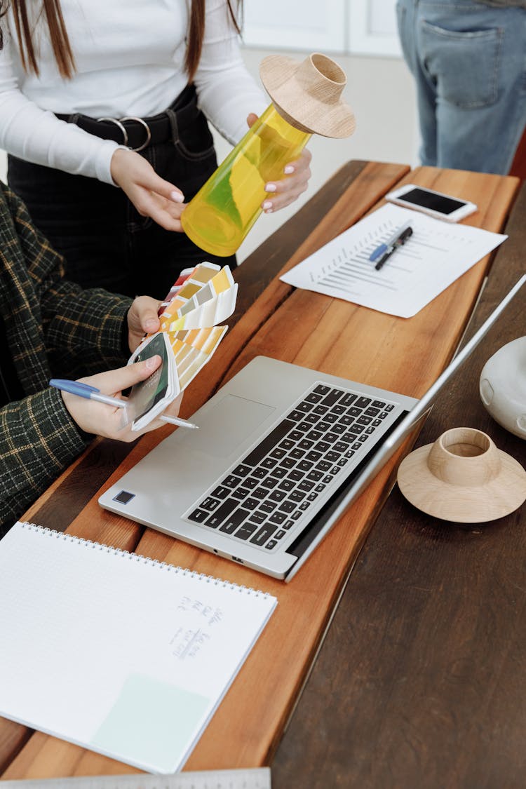 Person Holding Color Swatches And Glass Vase Beside Laptop On Wooden Table