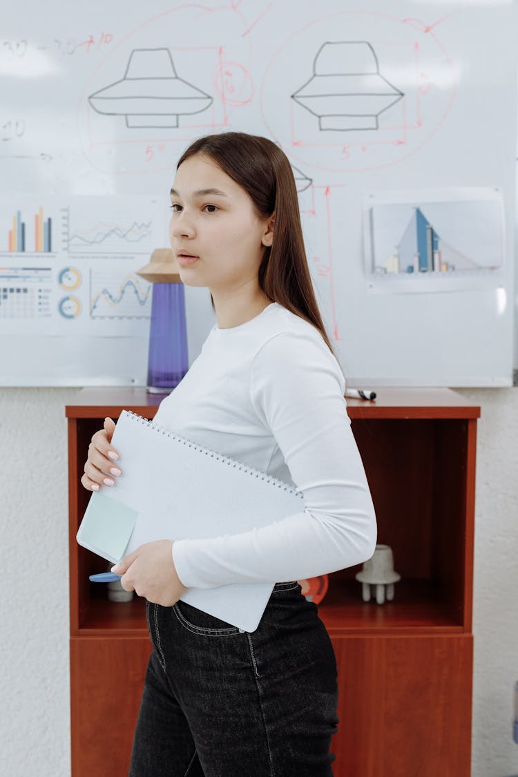 Young Woman In Black Jeans Holding Notebook While Standing Beside Board With Sketch