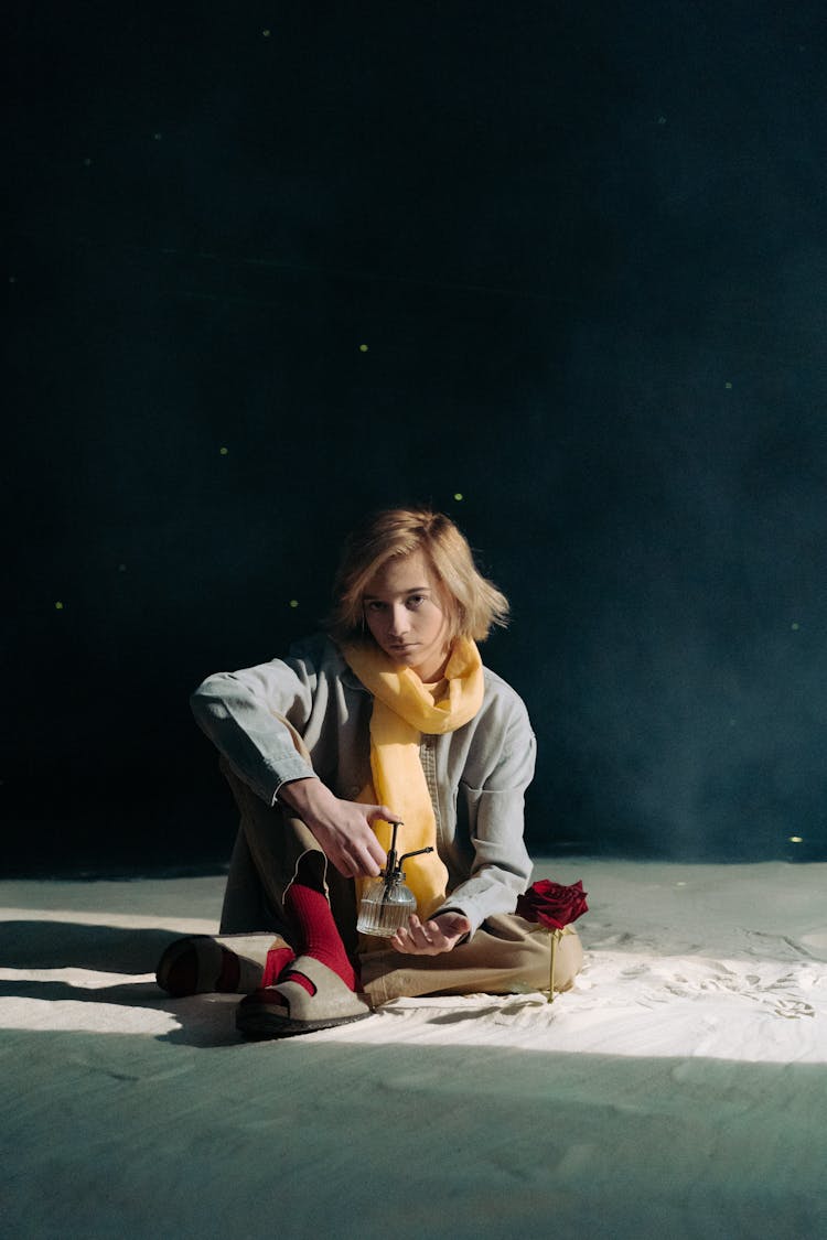 Boy With Yellow Scarf And Gray Blazer Sitting On White Sand Beside Red Rose