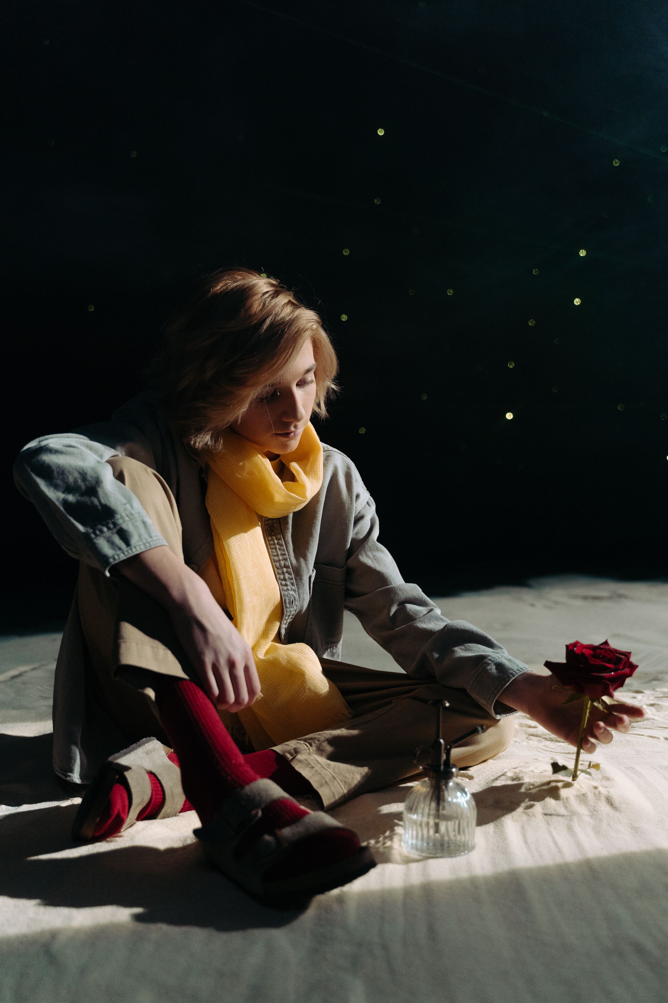 Person sitting on a floor holding a red rose, with a yellow scarf, captured in moody lighting.