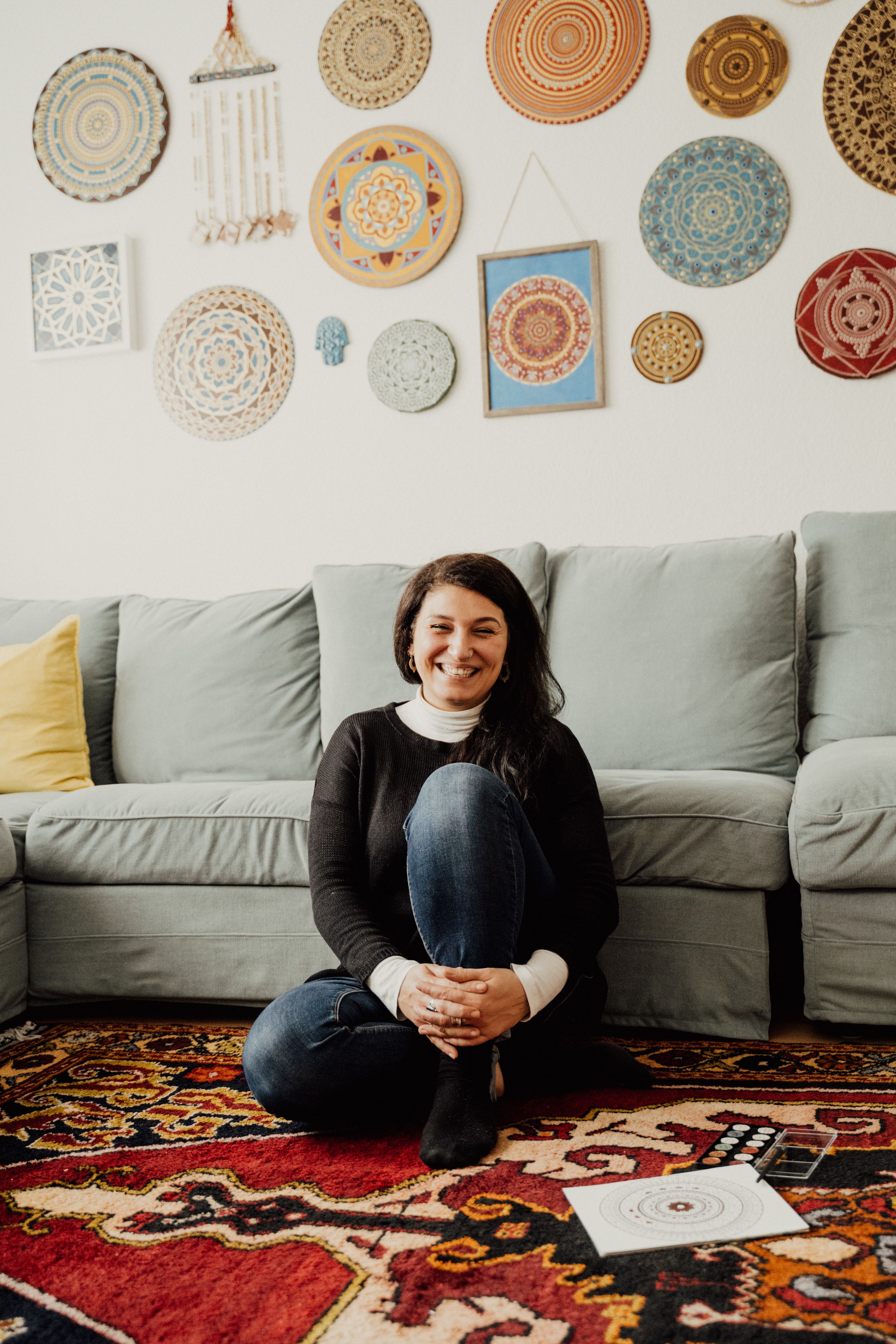 Portrait of a smiling woman sitting on a colorful rug in a decorated living room with mandalas on the wall.