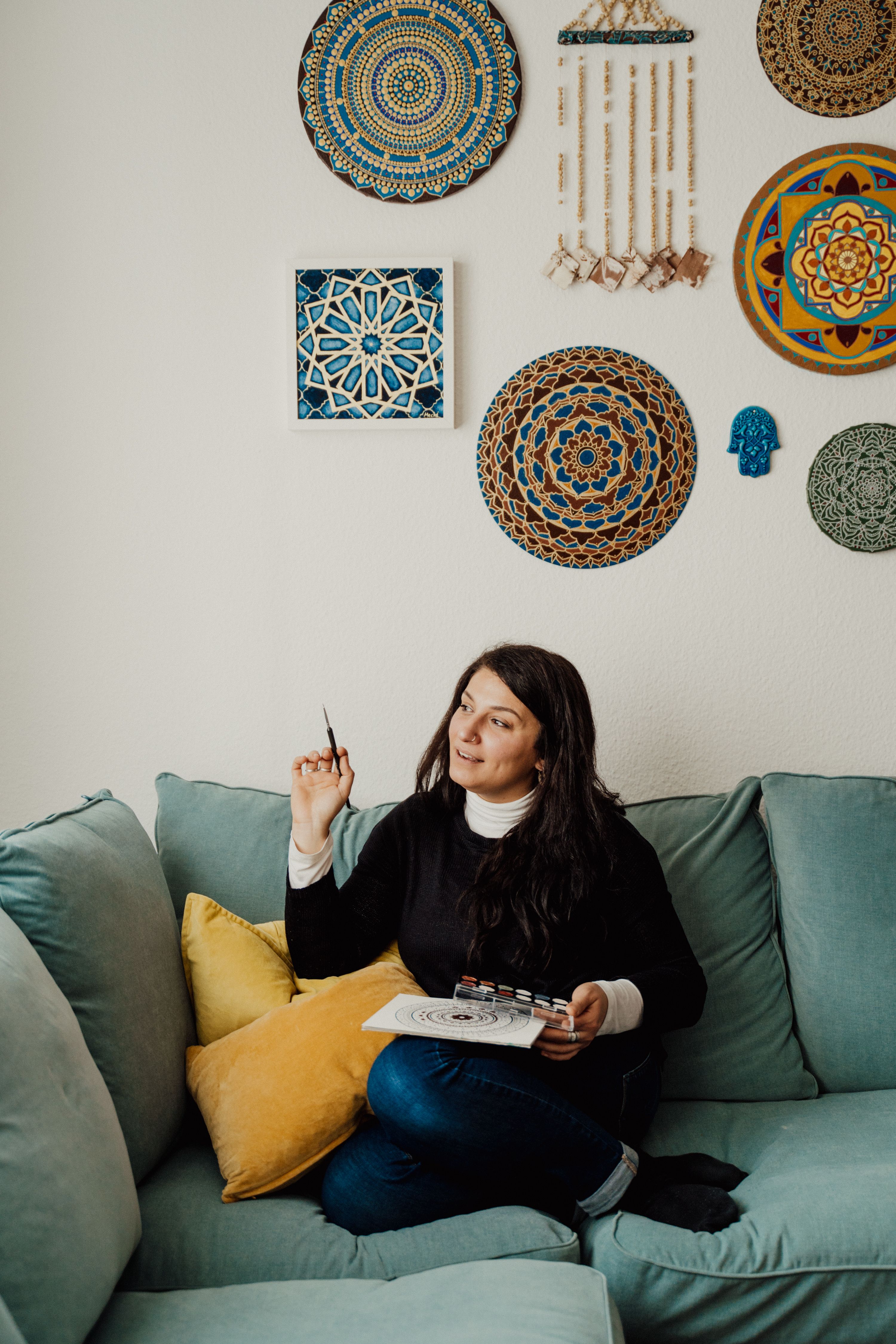 A thoughtful woman sits on a sofa with art supplies, contemplating a painting idea indoors.