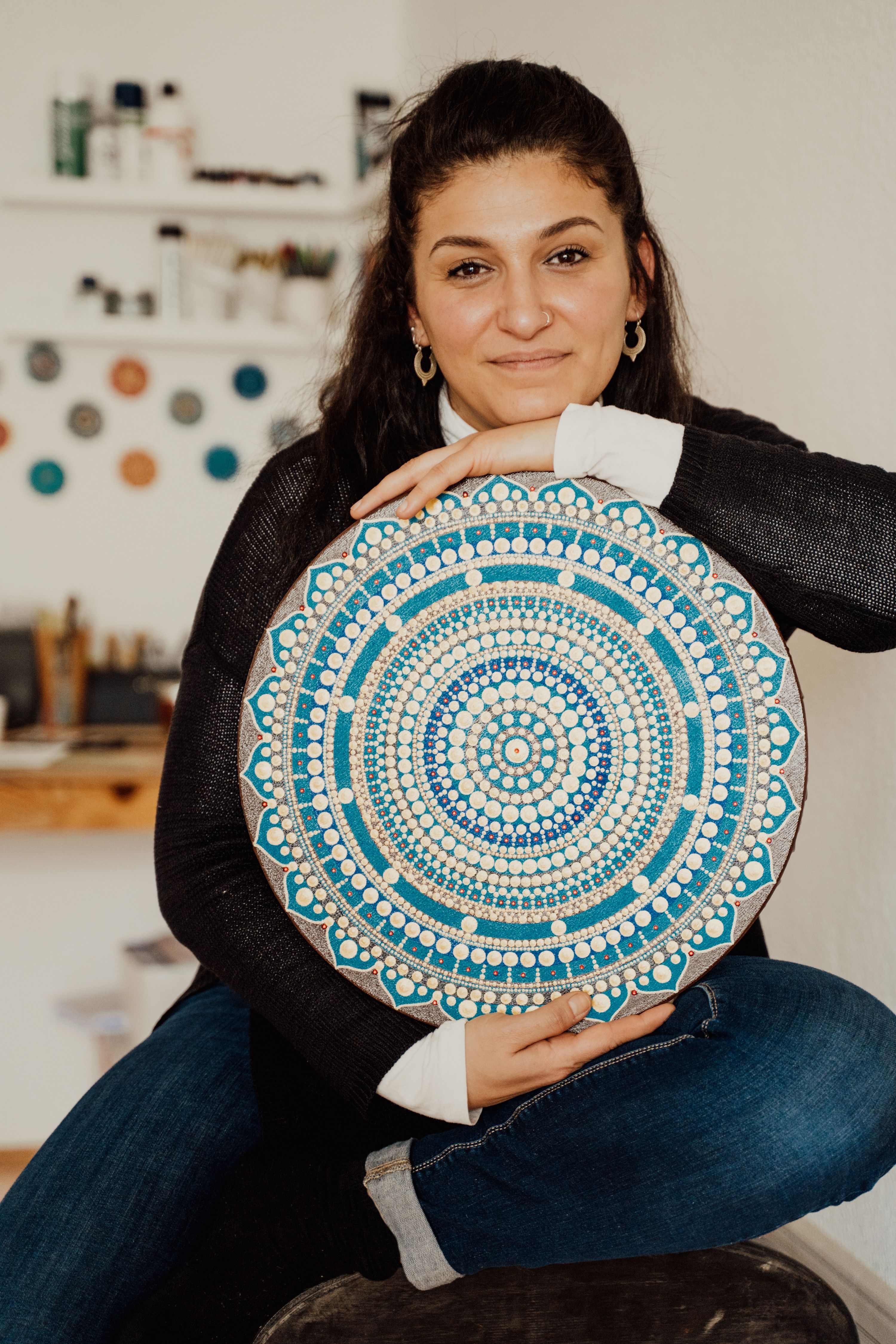Woman smiling and holding a handmade mandala painting indoors.