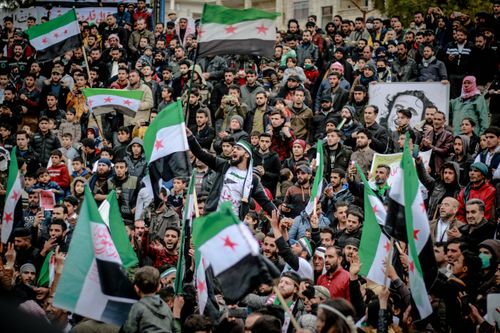 Free A large crowd gathers in a city square waving Syrian flags during a protest. Stock Photo