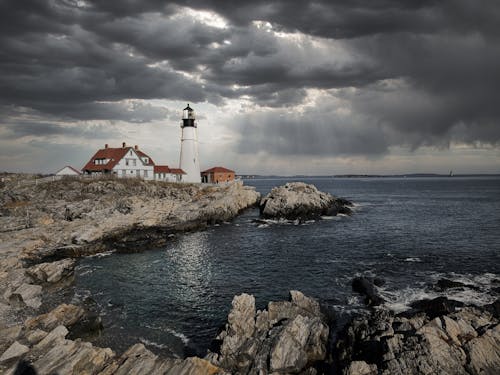 Amazing scenery of Portland Head Light lighthouse and small settlement located on rocky cliff above wavy sea under dramatic overcast sky