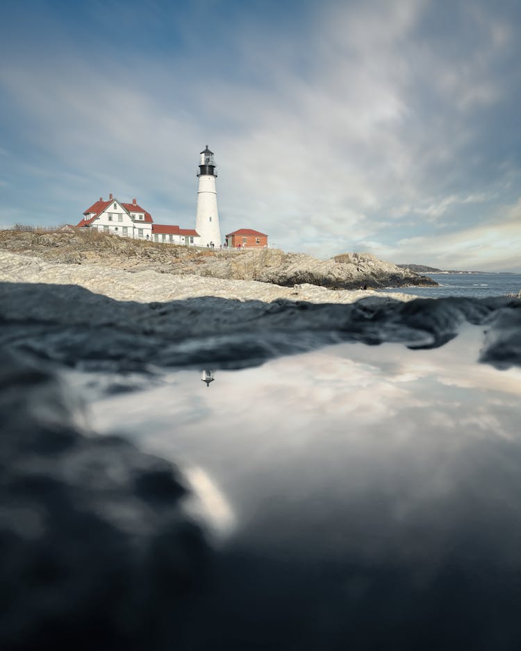 White Lighthouse And Cottages On Rocky Seashore Against Cloudy Sky