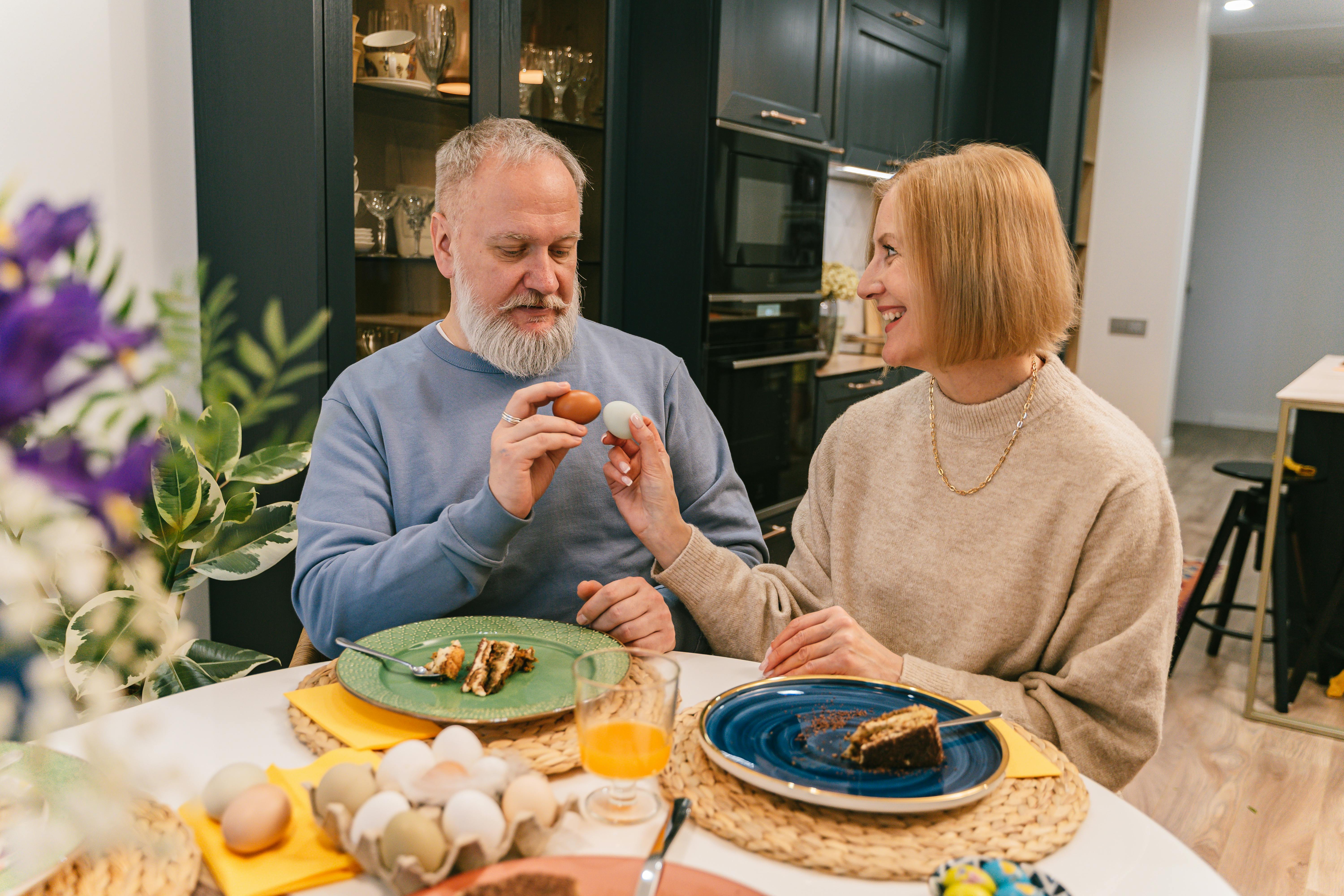 Free Elderly couple exchanging Easter eggs at a dining table, smiling warmly indoors. Stock Photo