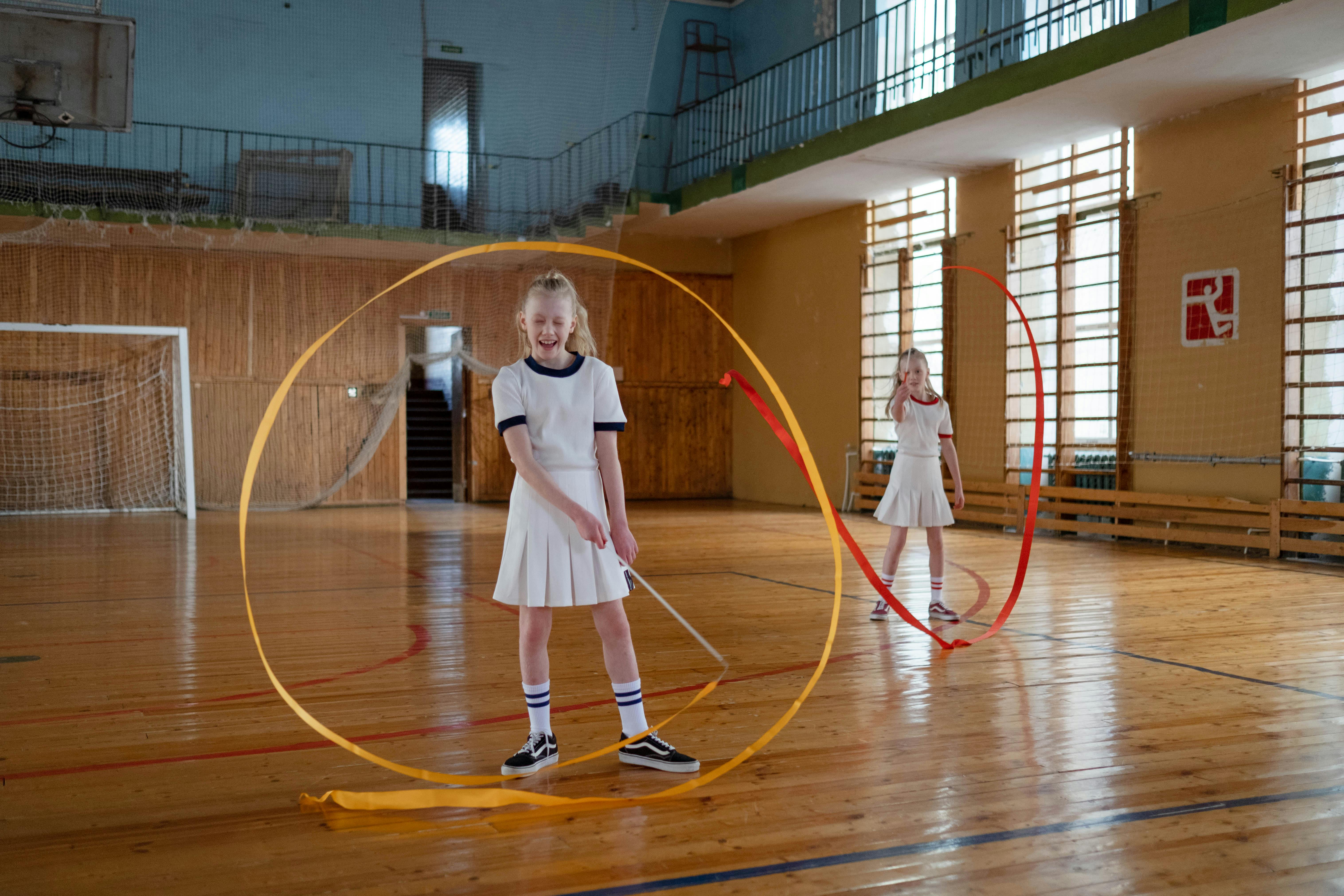Two young girls playfully practice rhythmic gymnastics with ribbons in a sports hall.