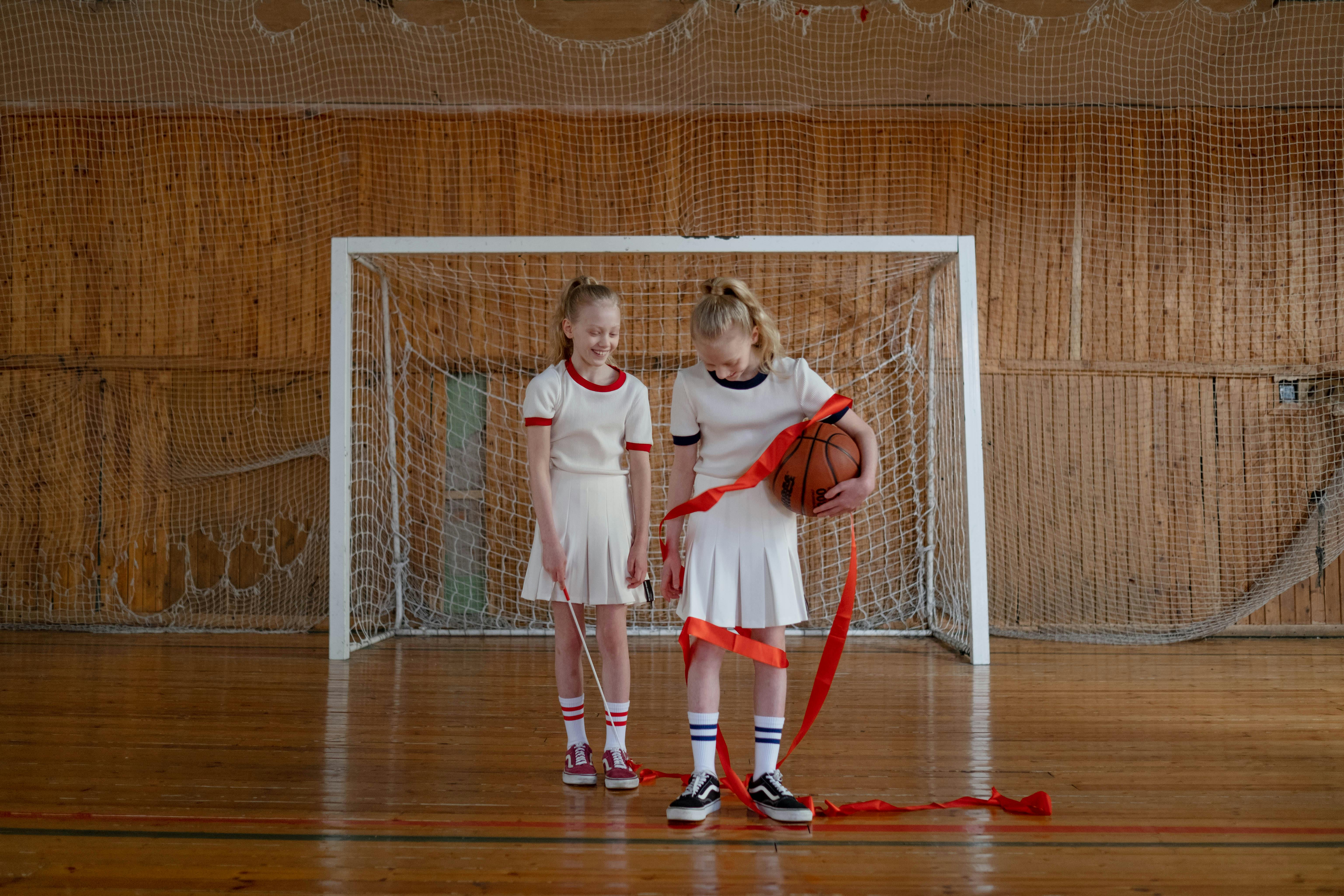 Two young girls standing in a gym celebrating with a basketball and ribbon.