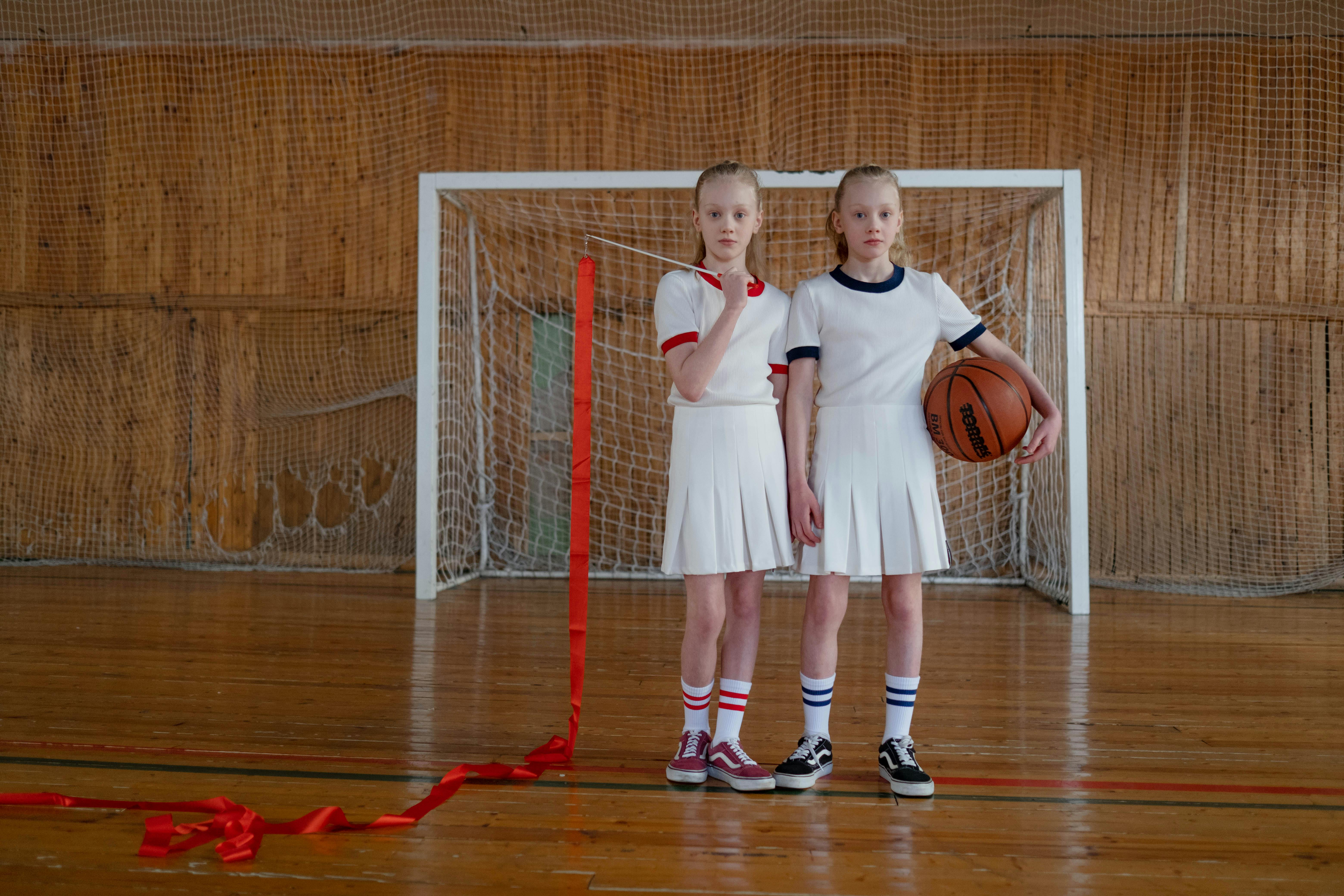 girls standing on the wooden floor holding a basketball and a red ribbon