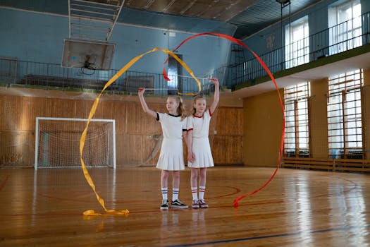 Two young girls performing rhythmic gymnastics with ribbons in an indoor gym.