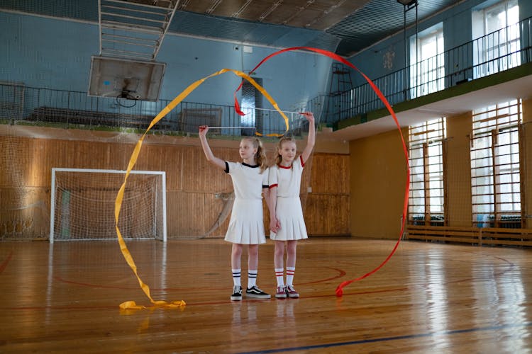 Twin Girls In White Dresses With Flowing Ribbons Standing On School Gym