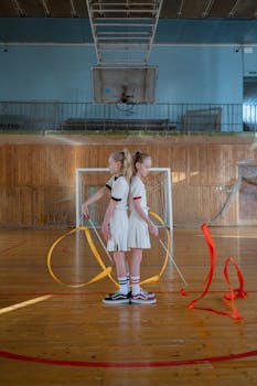 Two young gymnasts performing synchronized ribbon routines indoors on a sports court.