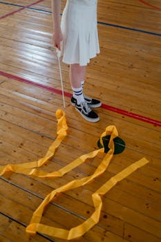 A young gymnast in sneakers holding a yellow ribbon on a wooden gym floor.