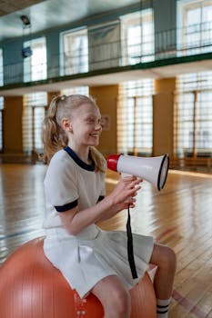 Young girl with a megaphone sitting on a fitness ball indoors, smiling and relaxed.