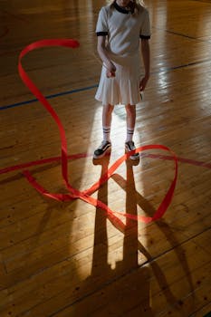 A young girl practicing rhythmic gymnastics with a red ribbon in a sunlit sports hall.