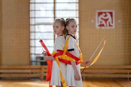 Young twin girls practicing rhythmic gymnastics with ribbons inside a sport hall.