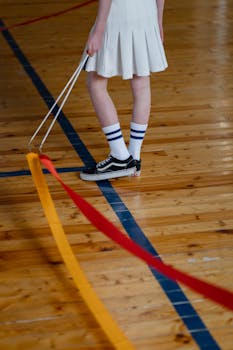 A child holding a gymnastics ribbon stands on a wooden floor in a gymnasium.