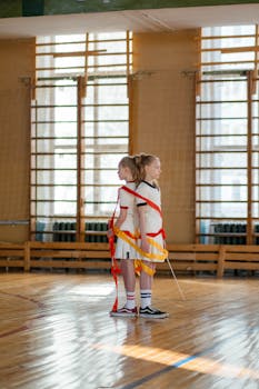 Two young girls wrapped in ribbons practicing rhythmic gymnastics in a sunlit school gym.