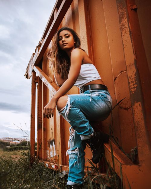 Free Fashionable woman in ripped jeans and white top posing against an orange container. Stock Photo