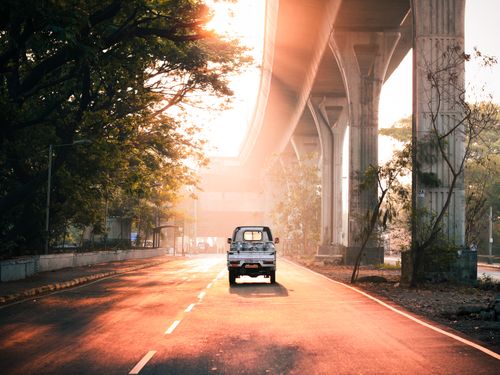 Free A lone vehicle drives on an urban street at sunrise, showcasing warm light and towering bridge. Stock Photo