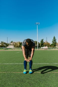 A male athlete in sportswear stretching on a football field under a clear blue sky.