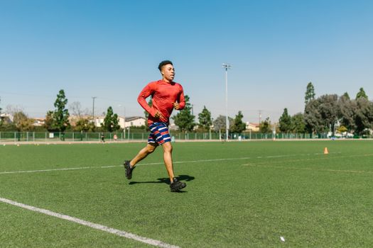A young man jogging on a sunny day on a sports field, wearing athletic gear.