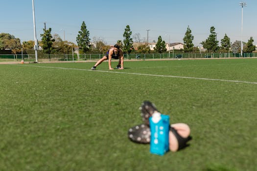 A focused Asian man stretches on a grassy field, ready for a workout, sports drink nearby.