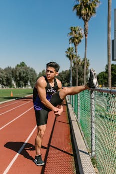 Young male athlete stretching on a sunny outdoor track with palm trees in the background.