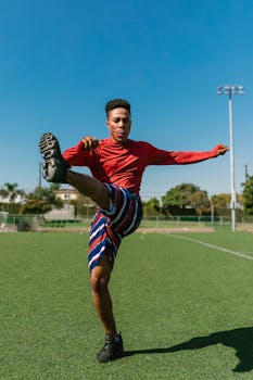 A young soccer player in sportswear stretching and warming up on a green field under a clear blue sky.