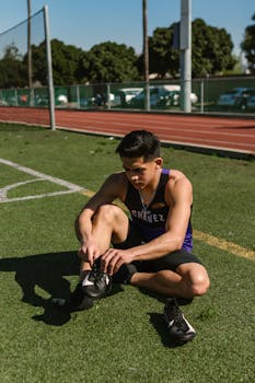 A young male athlete ties his shoes while preparing for a soccer game on a sunny outdoor field.