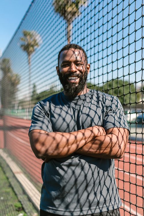 Free A cheerful man stands by a tennis court fence under bright sunlight, capturing a warm and lively outdoor moment. Stock Photo