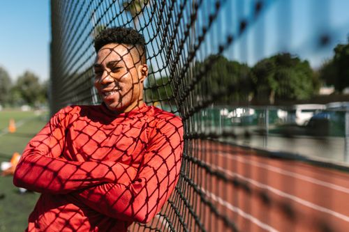 Free A cheerful athlete in a red shirt leaning on a sports field fence on a sunny day. Stock Photo