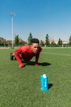 A young male athlete doing push-ups outdoors on a sunny day with a water bottle on the field.