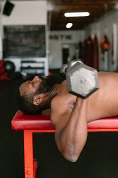 A muscular man exercises with dumbbells on a gym bench, showcasing strength and fitness.