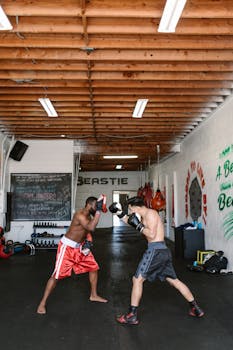 Two men engaged in a boxing match indoors with gym equipment surrounding them.