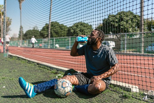 A Boy Sitting on a Green Field Near a Soccer Ball while Drinking Beverage