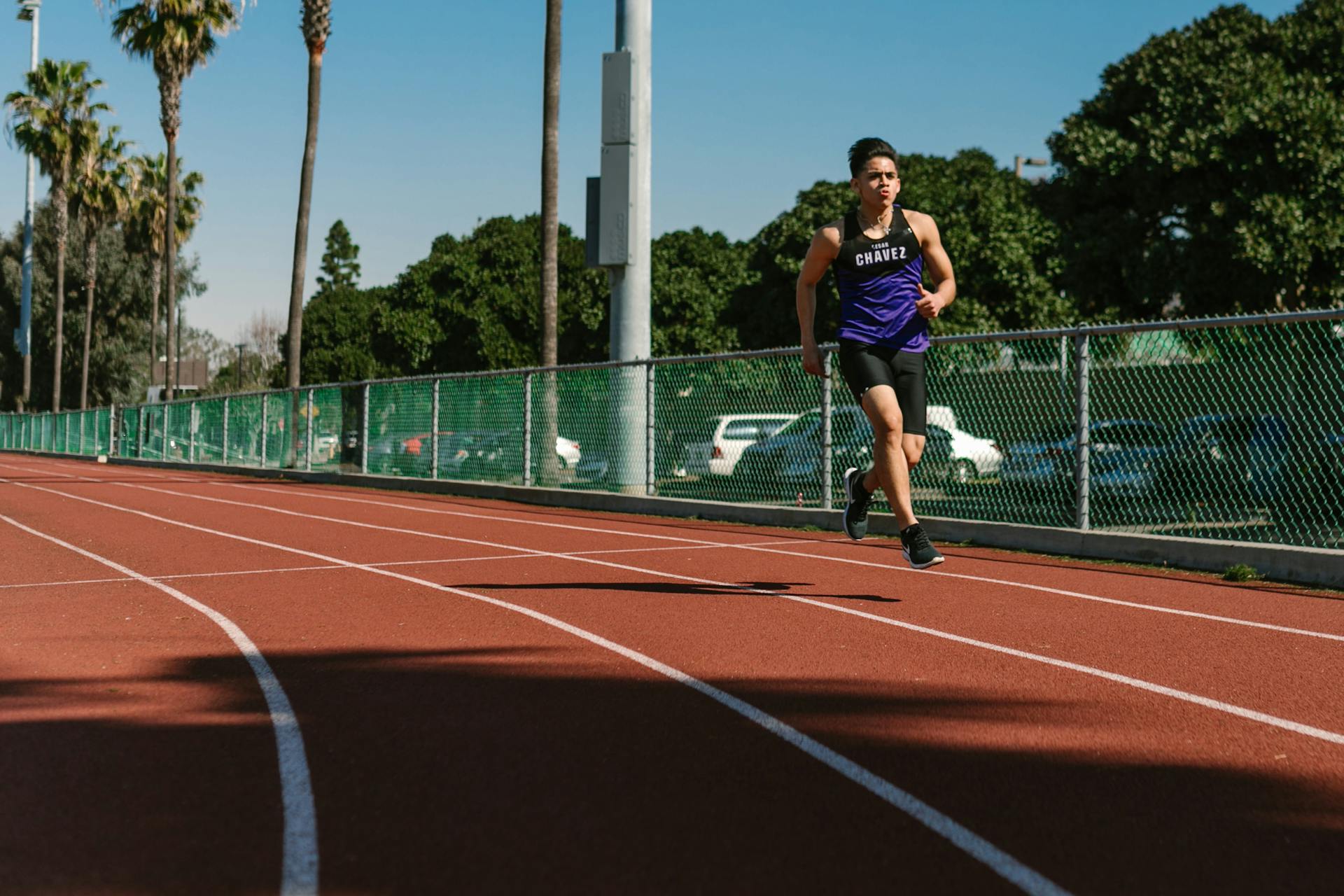 A young male athlete in a purple jersey running on an outdoor track with palm trees.