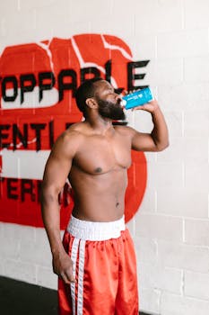 Shirtless muscular black man in gym shorts drinks water in front of motivational mural.