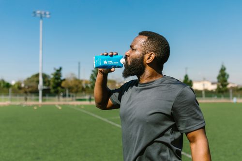 Free An adult man drinks a refreshing beverage outdoors on a sunny day, with a sports field in the background. Stock Photo