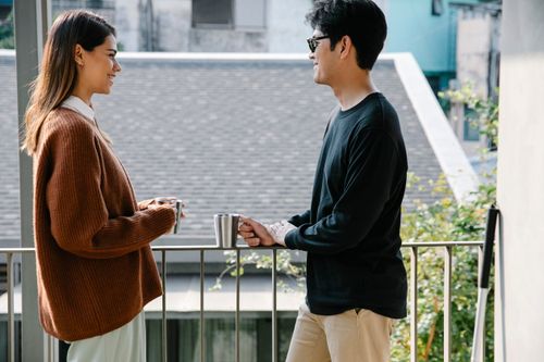 Free Two adults enjoying a relaxed conversation on a balcony, drinking coffee and smiling. Stock Photo