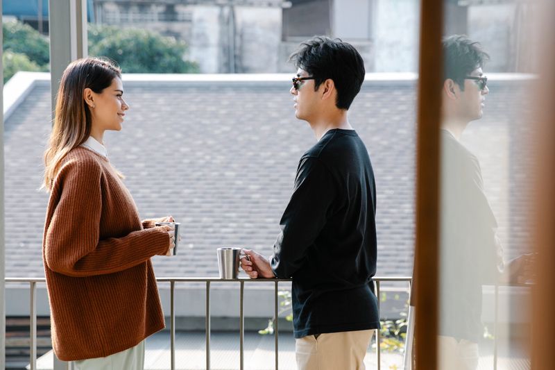 A man and woman enjoying a relaxed morning conversation with coffee on a sunny balcony.