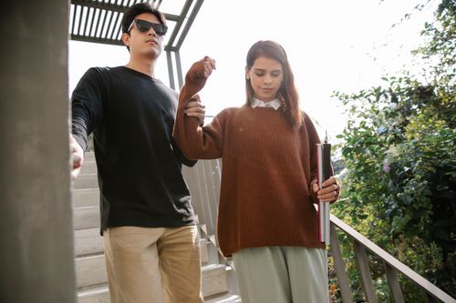 Free A man with sunglasses is guided by a woman with a cane while descending stairs outdoors. Stock Photo