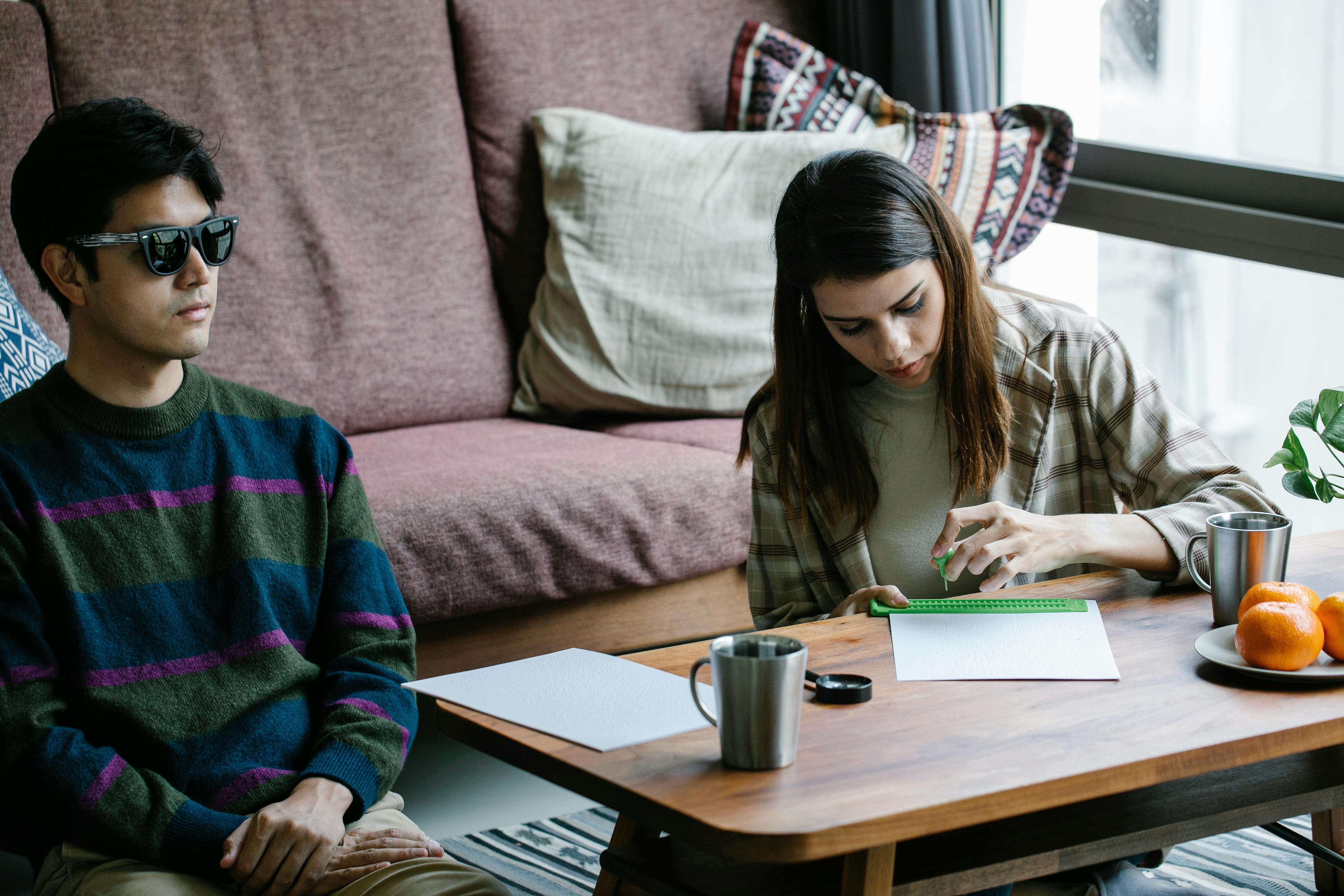A man with visual impairment relaxes as a woman writes in Braille at a home setting.