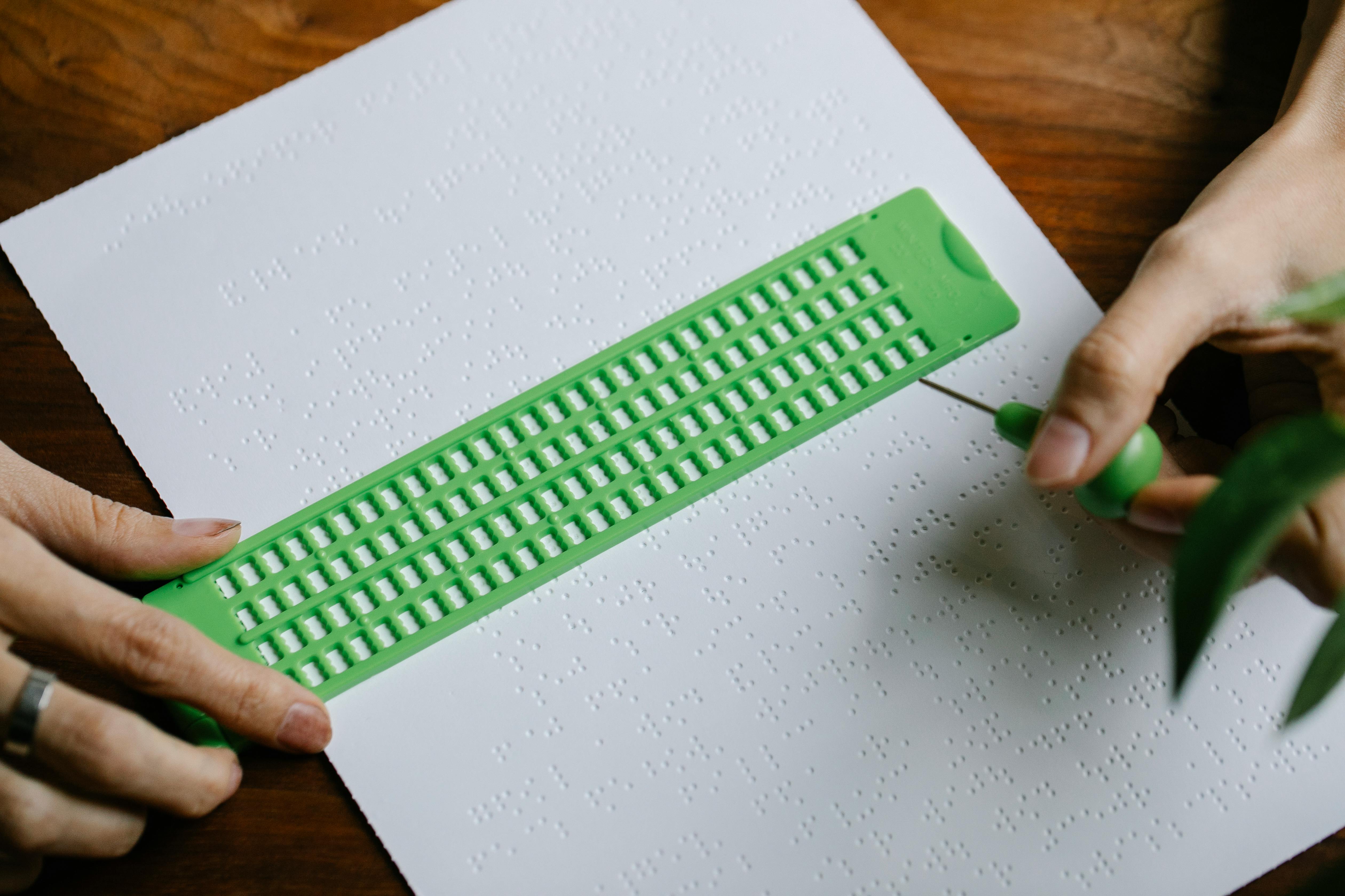 Close-up of hands using a green slate and stylus on Braille paper, emphasizing tactile writing.